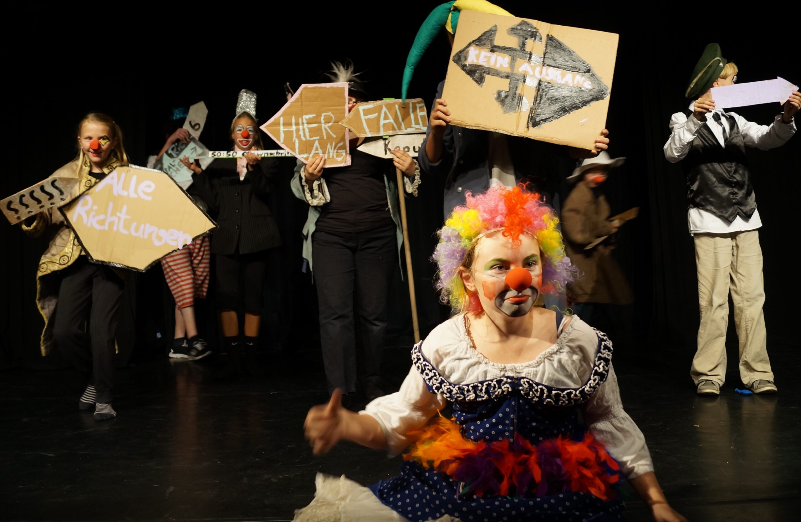 Ein Mädchen im Clownskostüm sitzt dem Publikum zugwendet auf dem Boden, im Hintergrund stehen Kinder, die Schilder hochhalten mit den Aufschriften "hier lang", "alle Richtungen", "Wegweiser" und "kein Ausgang"